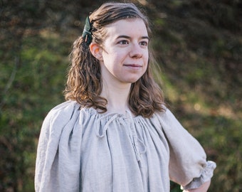May include: A woman wearing a light gray linen dress with a gathered neckline and puffed sleeves. She is standing in a grassy field and looking off to the side.