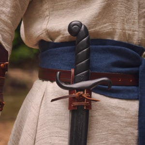 May include: A black sword with a curved guard and a spiral handle is sheathed in a brown leather scabbard. The sword is attached to a brown leather belt worn over a blue sash and a light-colored tunic. The sword's design suggests a historical or fantasy theme.