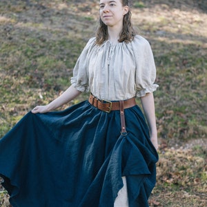 Pode incluir: Uma mulher vestindo uma blusa camponesa marrom claro com um cinto de couro marrom e uma saia azul escura. A saia &eacute; franzida na cintura e cai at&eacute; os tornozelos. A mulher est&aacute; em p&eacute; em um campo gramado.