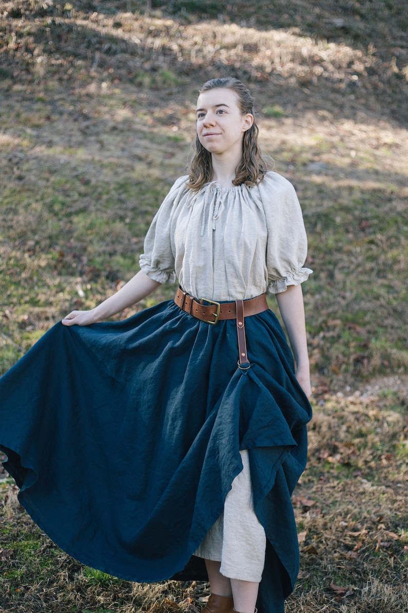 Pode incluir: Uma mulher vestindo uma blusa camponesa marrom claro com um cinto de couro marrom e uma saia azul escura. A saia &eacute; franzida na cintura e cai at&eacute; os tornozelos. A mulher est&aacute; em p&eacute; em um campo gramado.