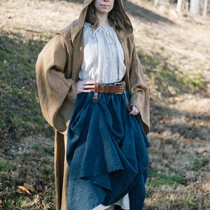 Pode incluir: Uma mulher vestindo uma t&uacute;nica marrom com capuz, uma saia azul e um cinto de couro marrom. Ela est&aacute; em p&eacute; em uma &aacute;rea gramada com &aacute;rvores ao fundo.