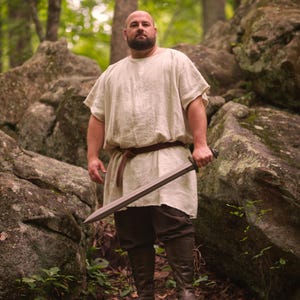 May include: A person wearing a cream-colored tunic, brown pants, and tall brown boots, holding a long sword. The person is standing in a forest setting with large rocks and green foliage. A brown belt is around the waist.