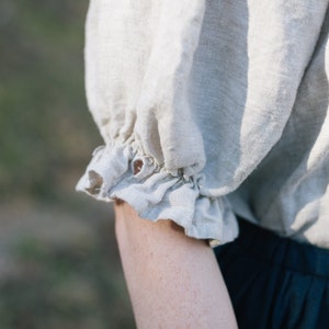 Pode incluir: Um close-up de uma camisa de linho bege claro com um detalhe de babado franzido na manga. A camisa &eacute; feita de um tecido macio e natural.