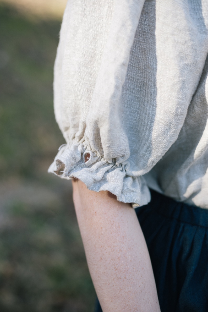 Pode incluir: Um close-up de uma camisa de linho bege claro com um detalhe de babado franzido na manga. A camisa &eacute; feita de um tecido macio e natural.