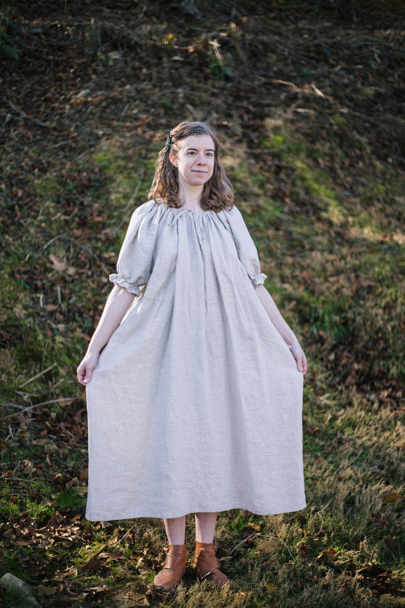 Pode incluir: Uma mulher vestindo um vestido longo de linho claro com decote drapeado e mangas curtas. Ela est&aacute; em p&eacute; em uma &aacute;rea gramada com folhas ca&iacute;das. Ela est&aacute; usando botas marrons.