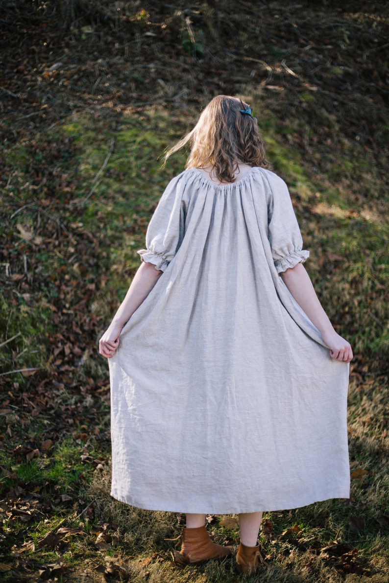 Pode incluir: Uma mulher vestindo um vestido longo de linho cinza claro com mangas bufantes. Ela est&aacute; em p&eacute; em uma &aacute;rea gramada com folhas marrons no ch&atilde;o.
