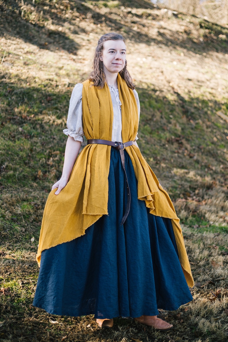 Pode incluir: Uma mulher vestindo uma saia longa azul escura com um xale amarelo mostarda drapeado sobre os ombros. Ela est&aacute; usando um cinto de couro marrom e tem o cabelo puxado para tr&aacute;s.