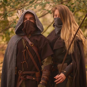 May include: Two people wearing hooded cloaks and face coverings stand in a forest. The person on the left is holding a bow and arrow. The person on the right is holding a bow.