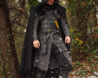 May include: A person wearing a medieval-style costume in a snowy forest. The outfit includes a black hooded cloak with fur trim, a leather cuirass, and matching boots. A yellow-handled dagger is visible at the waist.