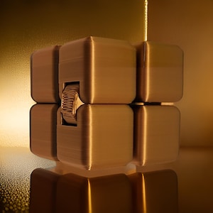 Puede incluir: Un juguete de fidget en forma de cubo de color dorado con una superficie texturizada. El juguete está formado por cubos más pequeños que se pueden girar y manipular.
