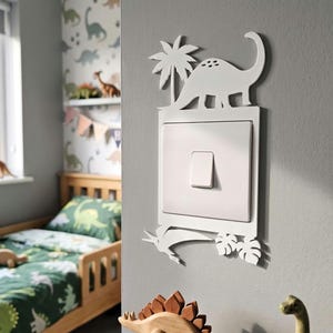 May include: A white light switch cover plate featuring a cutout design of a dinosaur and a palm tree at the top, and a pterodactyl with tropical leaves at the bottom. The square switch plate is mounted on a gray wall, surrounding a single rocker switch.