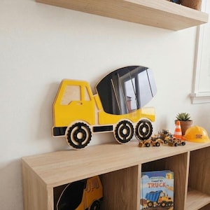May include: A yellow and black wooden cement mixer truck wall decoration featuring a layered design with a prominent rotating drum section, three circular wheels, and a cab, displayed as a flat, stylized construction vehicle cutout.