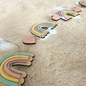 Puede incluir: Una guirnalda de luces LED con adornos de madera en forma de arcoíris y corazones alternados. Cada arcoíris consta de arcos superpuestos en amarillo pastel, rosa y turquesa. Los corazones son de color rosa pastel. Emiten luz blanca.
