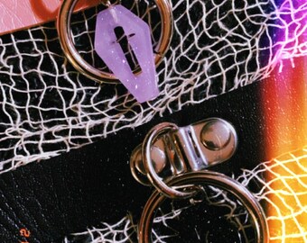 May include: Close-up of two chokers. One is pink, the other black. Each has a silver ring with a coffin-shaped charm. The charms are purple and black. The background is a white net over a black surface.