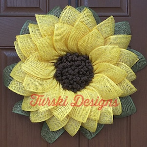 May include: A large yellow sunflower wreath with green burlap leaves. The sunflower is made of burlap and has a brown center.