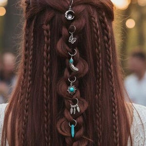 Op de afbeelding: Een persoon met lang, kastanjebruin haar draagt een witte kanten jurk met lange mouwen. Het haar is gestyled in een centrale vlecht, versierd met een verticale rij zilverkleurige haarbedels, waaronder een maan, vlinder, turkoois en veren.