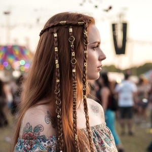 Op de afbeelding: Een vrouw met lang, roodbruin haar, gevlochten en versierd met gouden haaraccessoires. Ze draagt een off-the-shoulder top met bloemenprint. De afbeelding toont een bohemien of festival stijl.