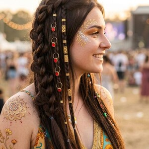 Könnte beinhalten: Eine Person mit langem braunem Haar, das zu einem dicken seitlichen Zopf geflochten ist, verziert mit kleinen goldenen Ringen mit roten kreisförmigen Akzenten und goldenen Haarspangen. Die Haut zeigt goldene temporäre Tattoos mit Blumen- und Sonnenmotiven.