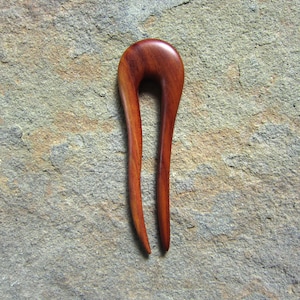 Può includere: Un fermaglio per capelli in legno con una finitura liscia di colore marrone rossiccio. Il fermaglio ha la forma di un anello curvo con un'estremità appuntita.