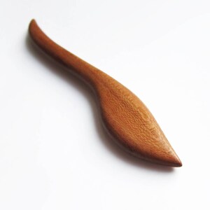 May include: A wooden hair stick with a curved, leaf-shaped design. The stick is a light brown color and has a smooth, polished finish.