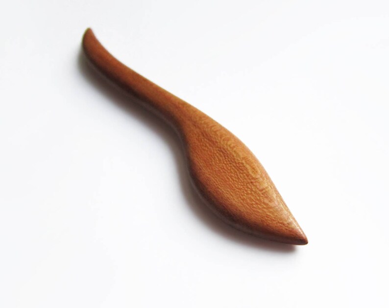 May include: A wooden hair stick with a curved, leaf-shaped design. The stick is a light brown color and has a smooth, polished finish.