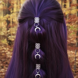 Peut inclure: Gros plan sur de longs cheveux violets profonds coiffés avec des accessoires argentés. Les cheveux sont ornés de pièces décoratives argentées avec des motifs géométriques et des breloques en forme de croissant de lune, créant une esthétique céleste.