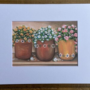 May include: Three terracotta flower pots with different colored flowers. The pots are decorated with white and blue dots. The flowers are yellow, white, and pink.