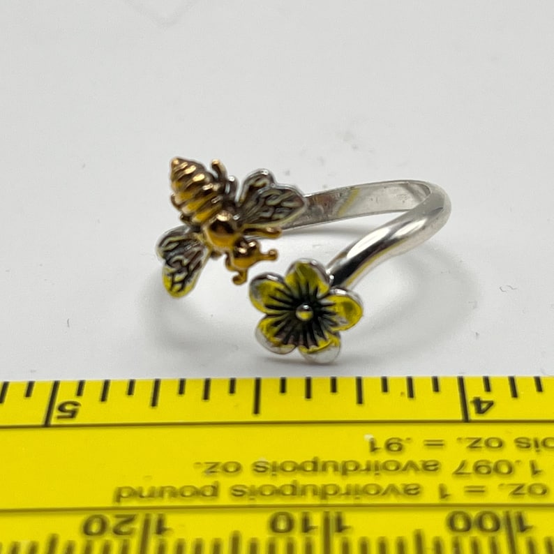 May include: A silver-toned ring featuring a detailed bee and a yellow flower. The bee has gold and black accents, while the flower has yellow petals and a black center. The ring is a nature-inspired jewelry piece.