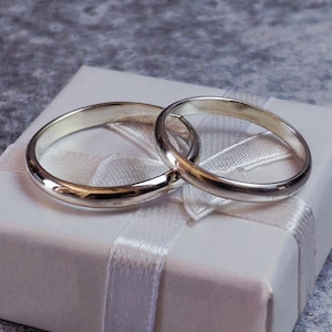 May include: Two silver wedding bands rest on a white gift box tied with a white satin ribbon. The rings are smooth and shiny, reflecting light. The box is square and the background is a textured gray surface.