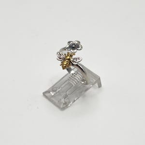 May include: A silver ring with a bee and flower design. The bee has gold-colored accents, and the flower is silver. The ring is displayed on a clear plastic stand against a white background.