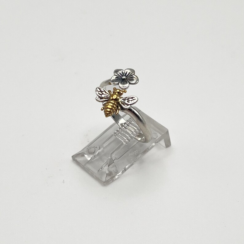 May include: A silver ring with a bee and flower design. The bee has gold-colored accents, and the flower is silver. The ring is displayed on a clear plastic stand against a white background.