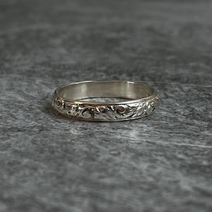 May include: A silver ring with an engraved floral pattern. The ring is circular and has a detailed design wrapping around the band. The ring is set against a gray background.