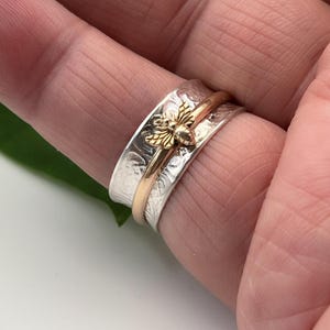 May include: A silver ring with a textured band and a gold bee charm. The ring has a gold band that spins freely. The bee charm is detailed with gold wings and body. The ring is shown on a finger.