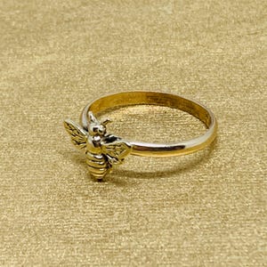 May include: A gold-toned metal ring featuring a small, detailed bee ornament attached to the front of the band. The bee has textured wings and a striped body, while the band itself is smooth and circular, creating a simple and cohesive design.