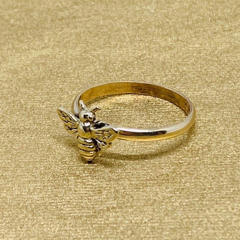 May include: A gold-toned metal ring featuring a small, detailed bee ornament attached to the front of the band. The bee has textured wings and a striped body, while the band itself is smooth and circular, creating a simple and cohesive design.
