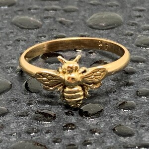 May include: A gold-toned ring featuring a detailed bee motif centered on the band. The bee has textured wings and a segmented body, with a smooth, polished finish on the circular band. The ring is crafted from a metallic material with a warm yellow hue.
