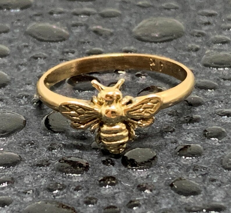 May include: A gold-toned ring featuring a detailed bee motif centered on the band. The bee has textured wings and a segmented body, with a smooth, polished finish on the circular band. The ring is crafted from a metallic material with a warm yellow hue.