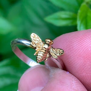 May include: A ring featuring a gold-toned bee ornament centered on a polished silver-toned band. The bee has detailed wings and a segmented body, contrasting with the smooth, reflective surface of the metallic band, creating a distinct visual focus.