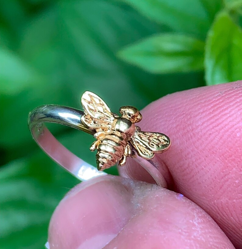 May include: A ring featuring a gold-toned bee ornament centered on a polished silver-toned band. The bee has detailed wings and a segmented body, contrasting with the smooth, reflective surface of the metallic band, creating a distinct visual focus.