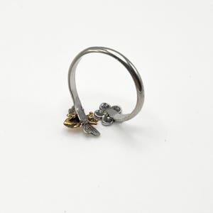 May include: A silver-toned ring with a bee and flower design. The bee has gold-colored wings and a black body. The flower has three petals. The ring is on a white background.