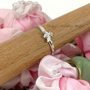 May include: A silver ring with a bee design. The ring is on a wooden dowel, surrounded by pink and green fabric. The bee has detailed wings and body. The ring is a delicate piece of jewelry.