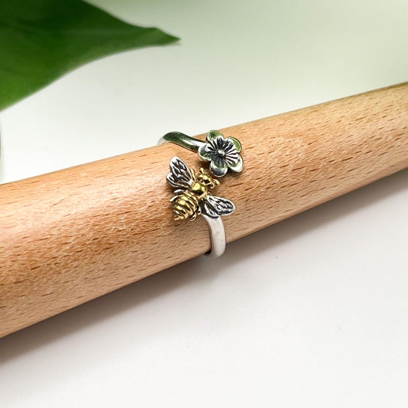 May include: A silver ring with a bee and flower design. The bee has gold-colored accents on its body and wings. The flower is silver with a dark center. The ring is displayed on a wooden dowel.