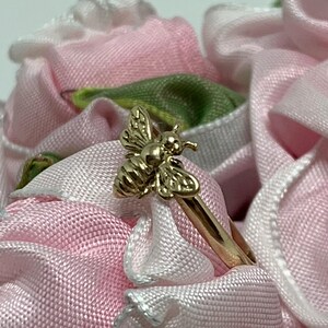 May include: A gold-toned ring featuring a detailed bee motif on the band. The bee is rendered with textured wings and a smooth, rounded body. The ring is positioned amidst soft, light pink fabric petals with subtle white trim and green accents.
