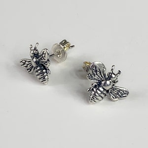 May include: A pair of silver-colored bee stud earrings. The earrings are detailed with textured wings and body segments, and have a post and butterfly backing. The earrings are displayed on a white background.