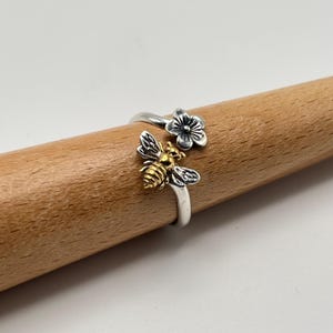 May include: A silver-colored ring with a bee and flower design. The bee has gold-colored accents and detailed wings. The flower is silver with dark detailing. The ring is wrapped around a wooden dowel.