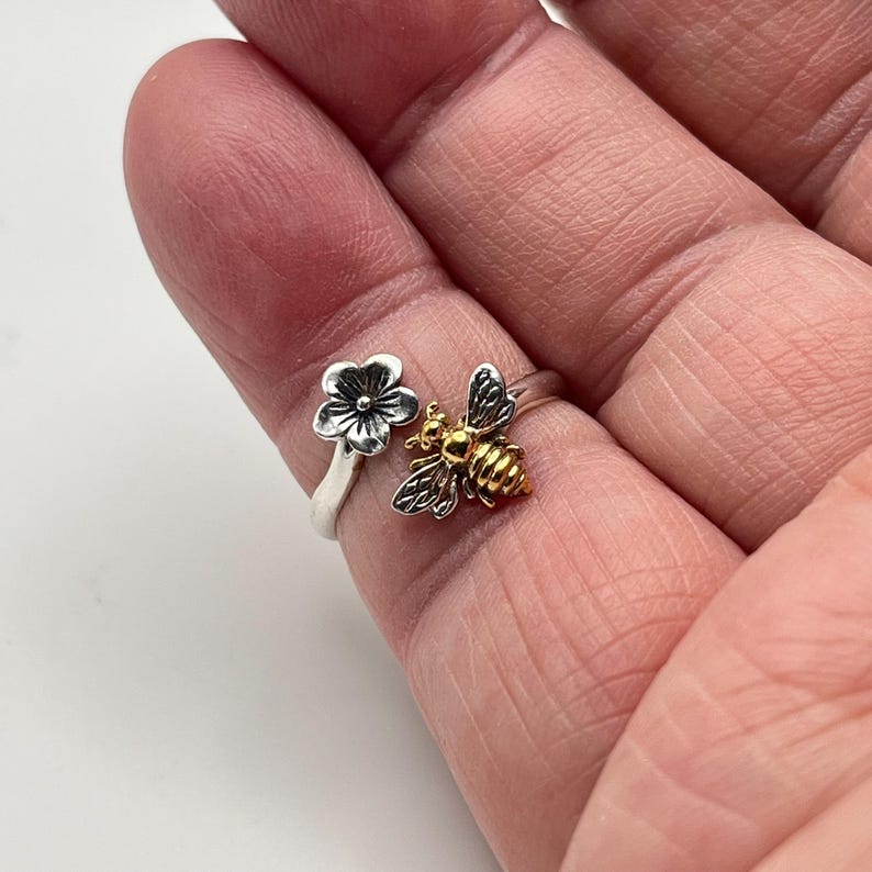 May include: A silver ring with a flower and bee design. The flower is silver with a black center, and the bee has gold-colored accents. The ring is on a finger.