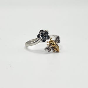 May include: A silver-toned ring featuring a flower and a bee design. The flower has a dark finish, while the bee is gold-toned with detailed wings. The ring is set against a white background.