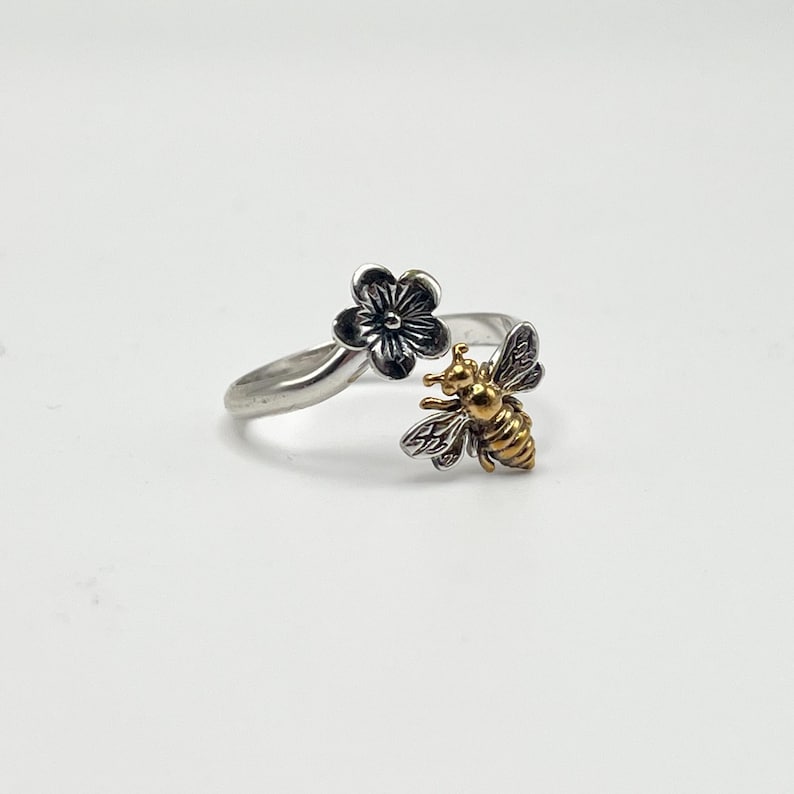 May include: A silver-toned ring featuring a flower and a bee design. The flower has a dark finish, while the bee is gold-toned with detailed wings. The ring is set against a white background.
