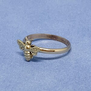 May include: A gold-toned metal ring featuring a small, detailed bee ornament attached to the front of the band. The bee has textured wings and a segmented body, while the band itself is smooth, polished, and circular in its overall design.