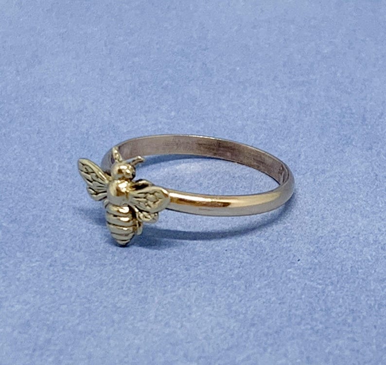 May include: A gold-toned metal ring featuring a small, detailed bee ornament attached to the front of the band. The bee has textured wings and a segmented body, while the band itself is smooth, polished, and circular in its overall design.